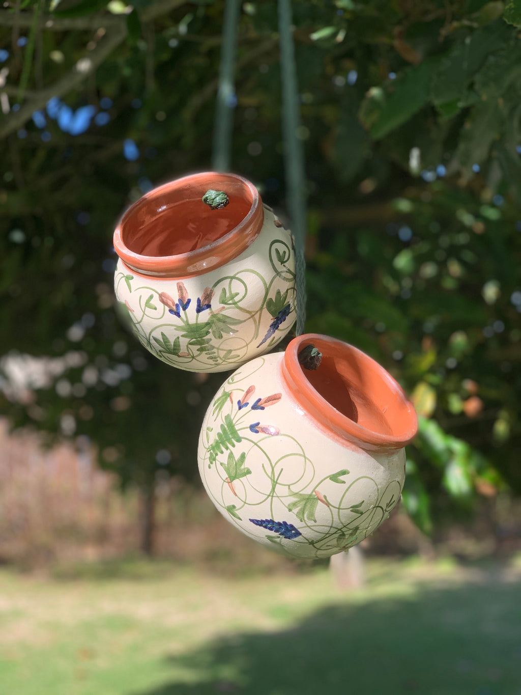 Two small hanging ceramic planters with terracotta-colored rims and green floral patterns, suspended outdoors against a blurred background of green foliage and grass.