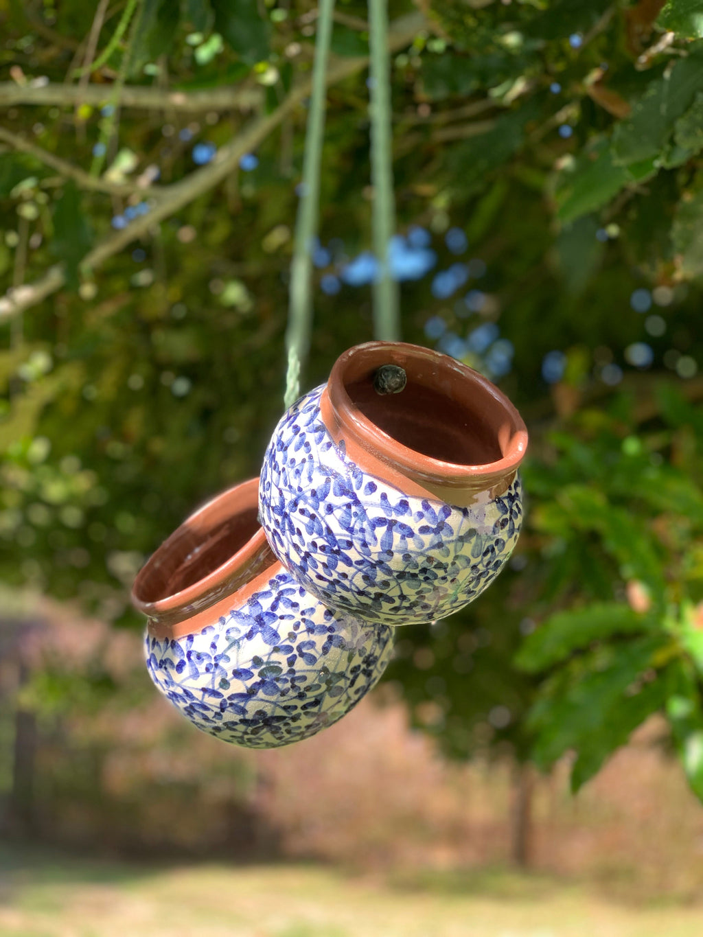 Two small hanging ceramic planters with blue floral patterns and terracotta rims, suspended by strings outdoors against a blurred green garden background.