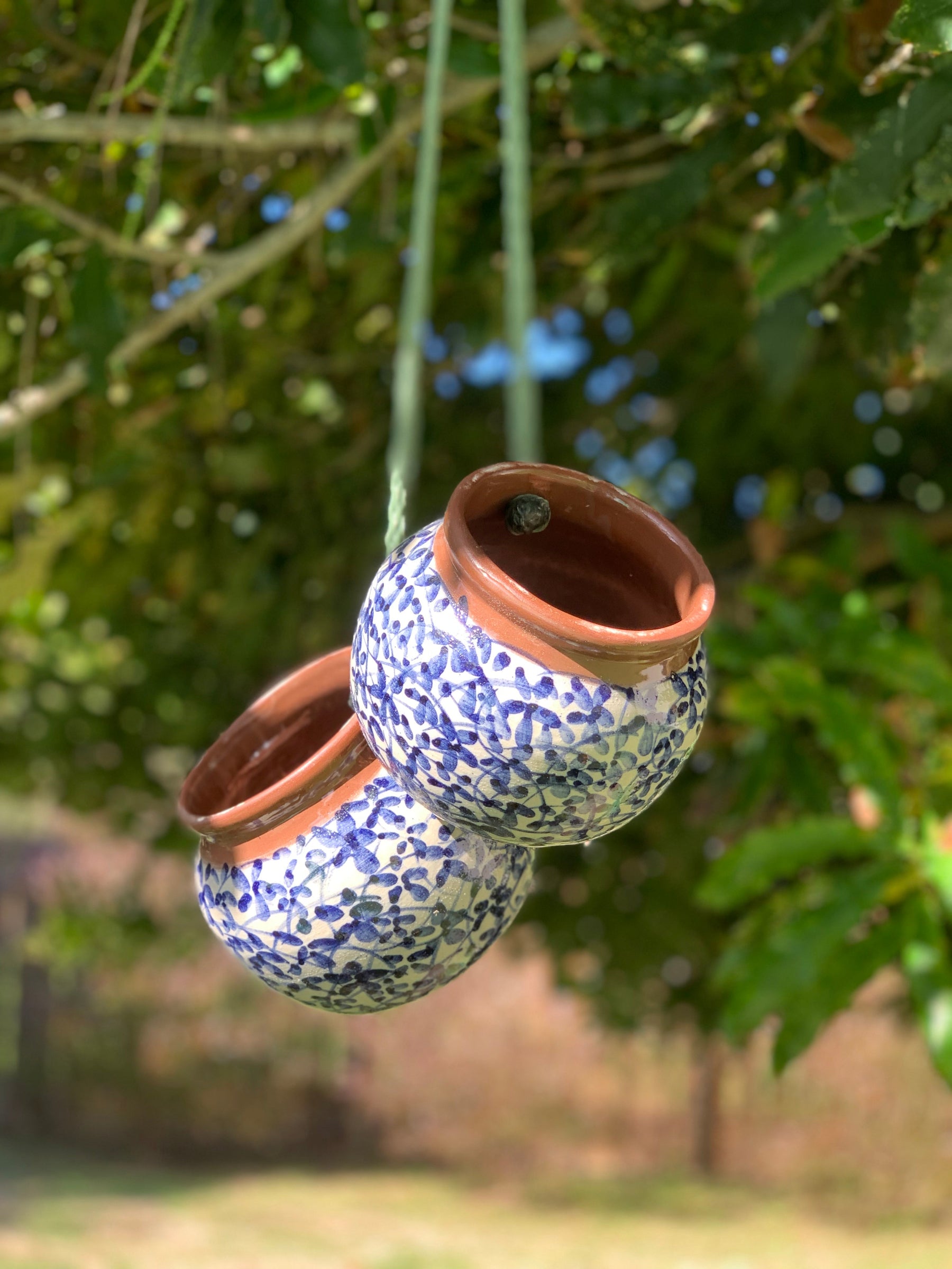 Two small hanging ceramic planters with blue floral patterns and terracotta rims, suspended by strings outdoors against a blurred green garden background.
