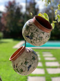 Two hanging ceramic planters with a cream glaze, brown terracotta rims, and green floral scrollwork, suspended outdoors in front of a blurred garden background.