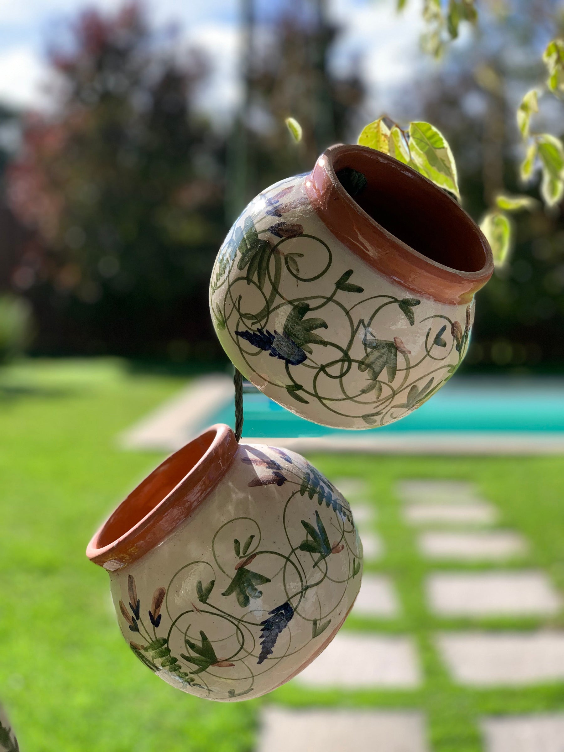Two hanging ceramic planters with a cream glaze, brown terracotta rims, and green floral scrollwork, suspended outdoors in front of a blurred garden background.