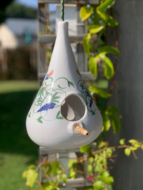 A white ceramic bird feeder with a teardrop shape, decorated with colorful painted birds, flowers, and vines. It hangs from a green string outdoors, with a wooden perch.