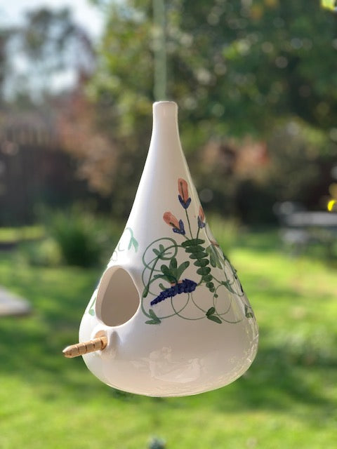 A white ceramic bird feeder with a teardrop shape, decorated with green vines and blue-purple flowers, hanging outdoors against a blurred green garden background.