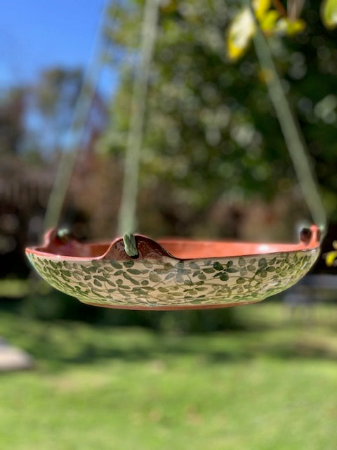 A hanging ceramic garden tray with a floral pattern and a terracotta-colored rim, suspended by cords in an outdoor garden setting.