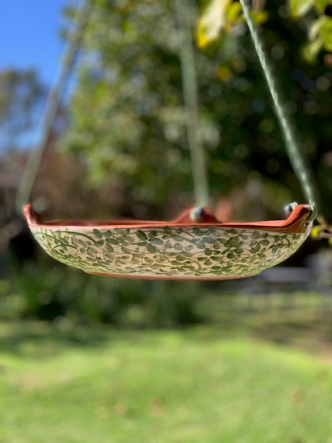 A hanging ceramic garden tray with a  green pattern and terracotta-colored rim, suspended by a green cord in an outdoor garden setting with a blurred background of grass, trees, and blue sky.